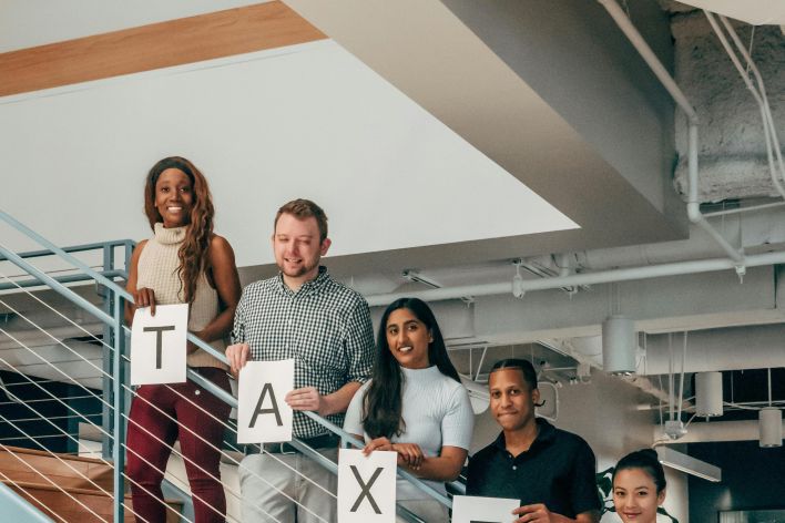 A group of diverse colleagues holding 'TAXES' sign while standing on a modern office staircase.